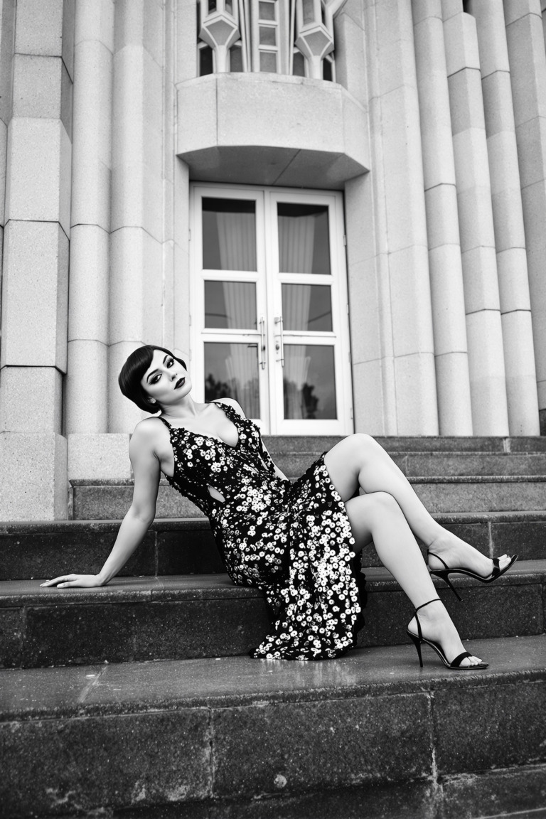 CC0. Public domain image. A woman lounges elegantly on the steps of an art deco building, wearing a shimmering flapper-style dress adorned with sequins. The monochromatic black-and-white image captures the sparkling details of her dress and the soft glow of her skin. Her short, bobbed hair frames her face, and her dark eyeliner adds a vintage charm. The geometric architecture of the background complements her striking pose, creating an atmosphere of sophistication and timeless glamour.
